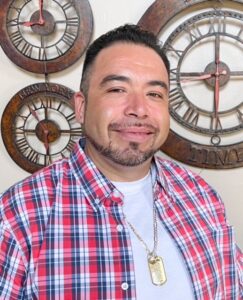 A man smiling in front of a wall with decorative clocks.